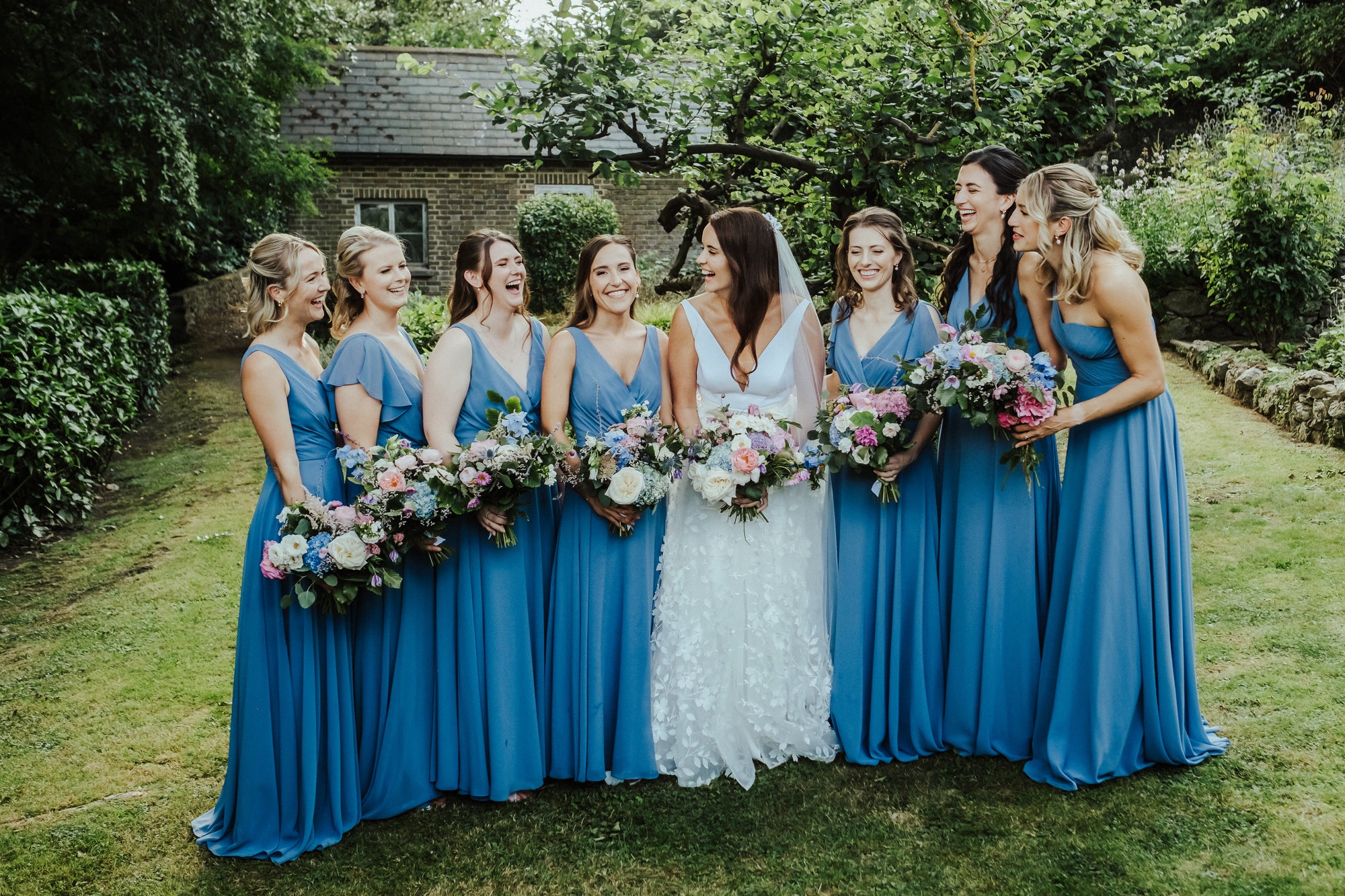 Bride with bridesmaids in blue dresses holding bouquets outdoors.