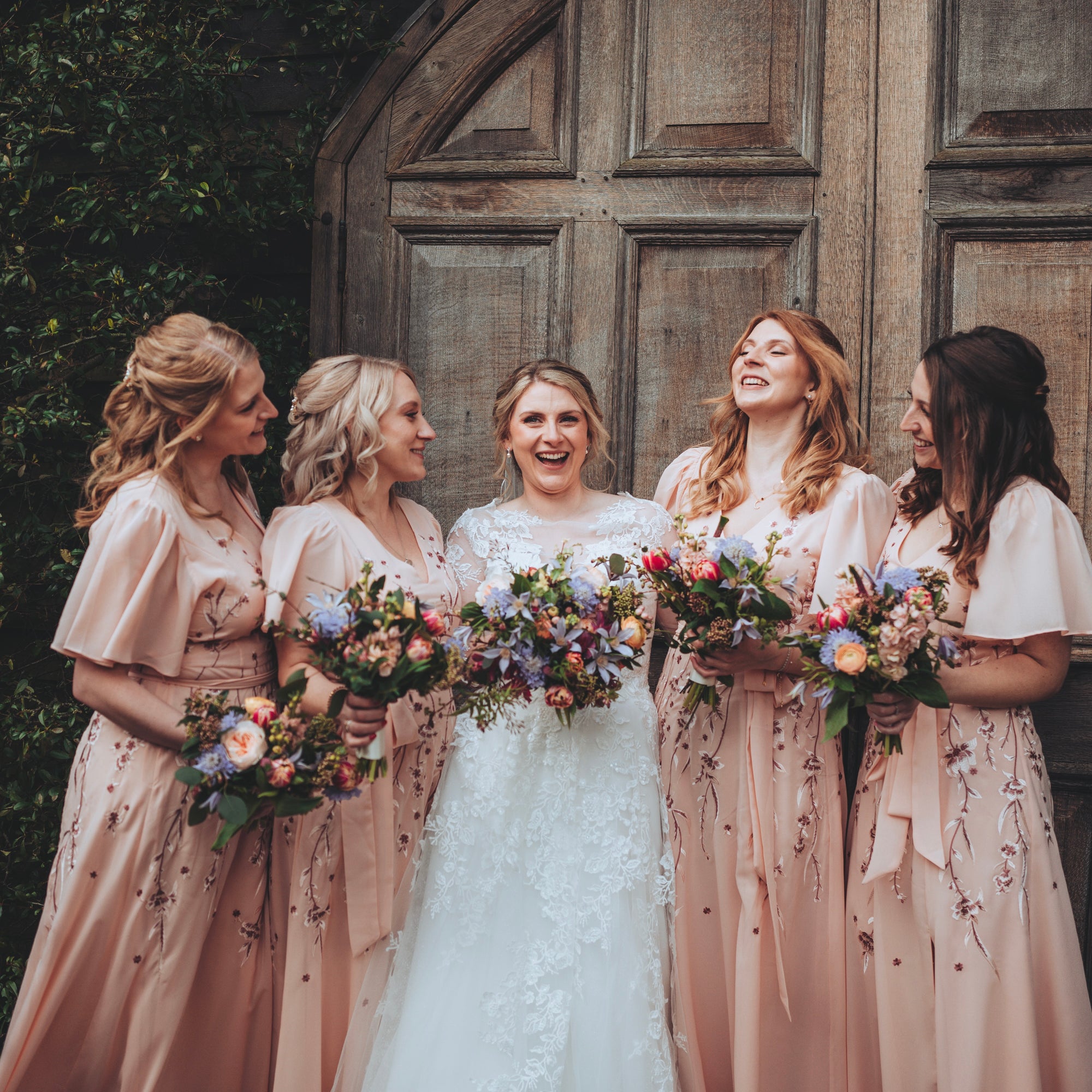 Bride with bridesmaids in matching dresses holding floral bouquets in front of a wooden door.